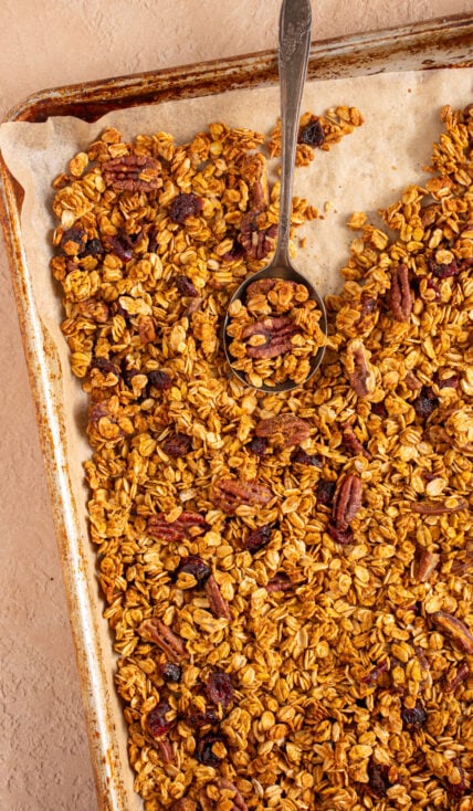 Sheet pan with parchment paper filled with pumpkin spice granola with dried cranberries and pecans with a spoon scooping some granola.