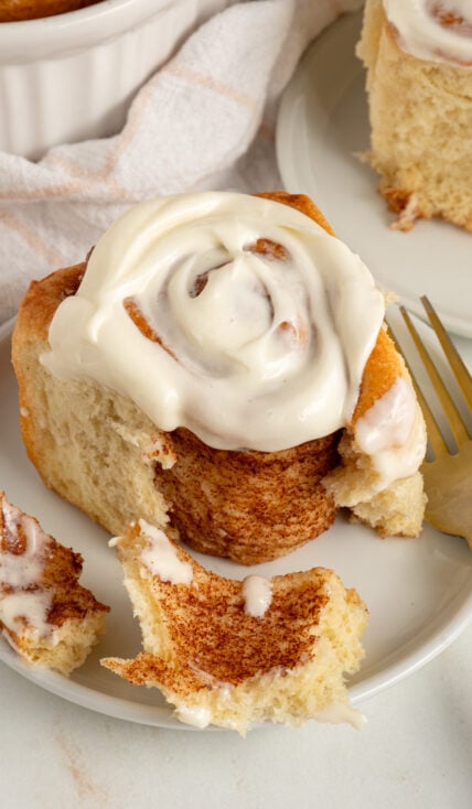 A fluffy cinnamon roll on a plate covered in cream cheese frosting and opened up slightly to reveal the cinnamon insides.